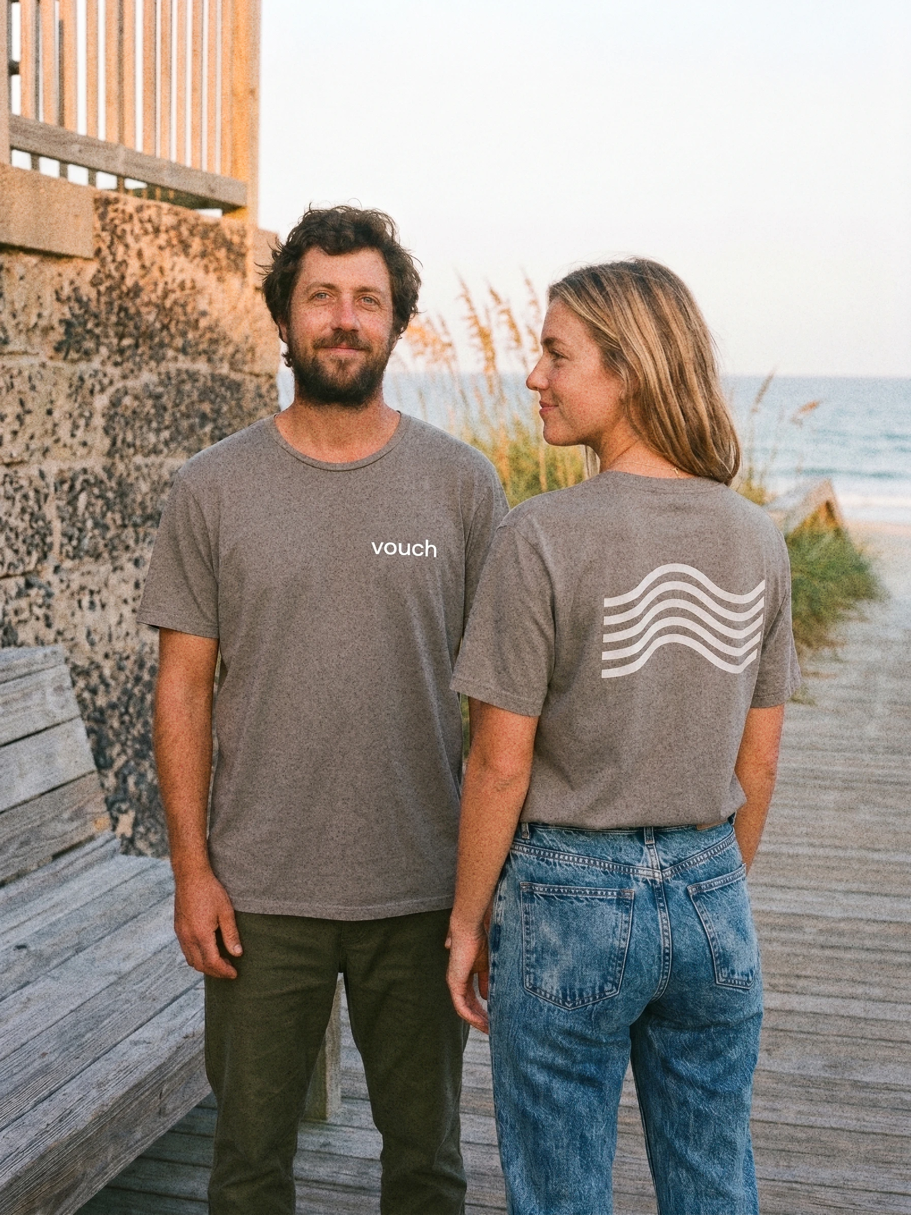 Two people wearing grey Vouch-branded t-shirts on a beach boardwalk, showing the front wordmark and back wave graphic